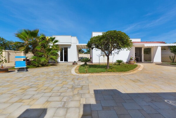 A courtyard with a paved walkway, palm trees, and a building with a white facade and red roof. - 2092 Villa Il Fenicottero Salentino (Manduria)