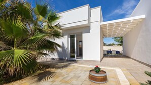 A modern building with a patio area featuring stone tiles, a wooden barrel planter, and palm trees. - 2092 Villa Il Fenicottero Salentino (Manduria)