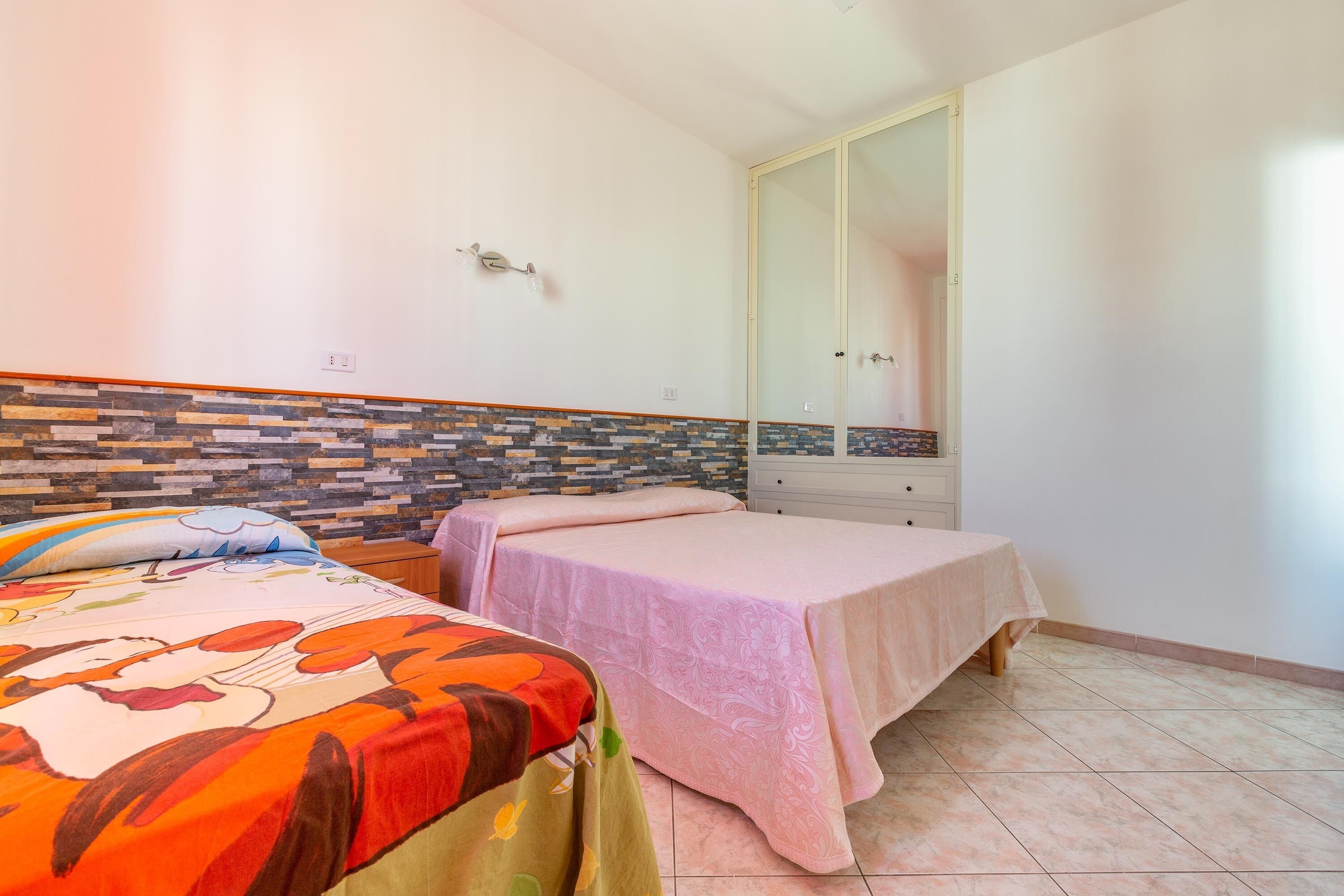 A bedroom with two beds, a tiled floor, a wooden headboard, a wardrobe, and a mirror.