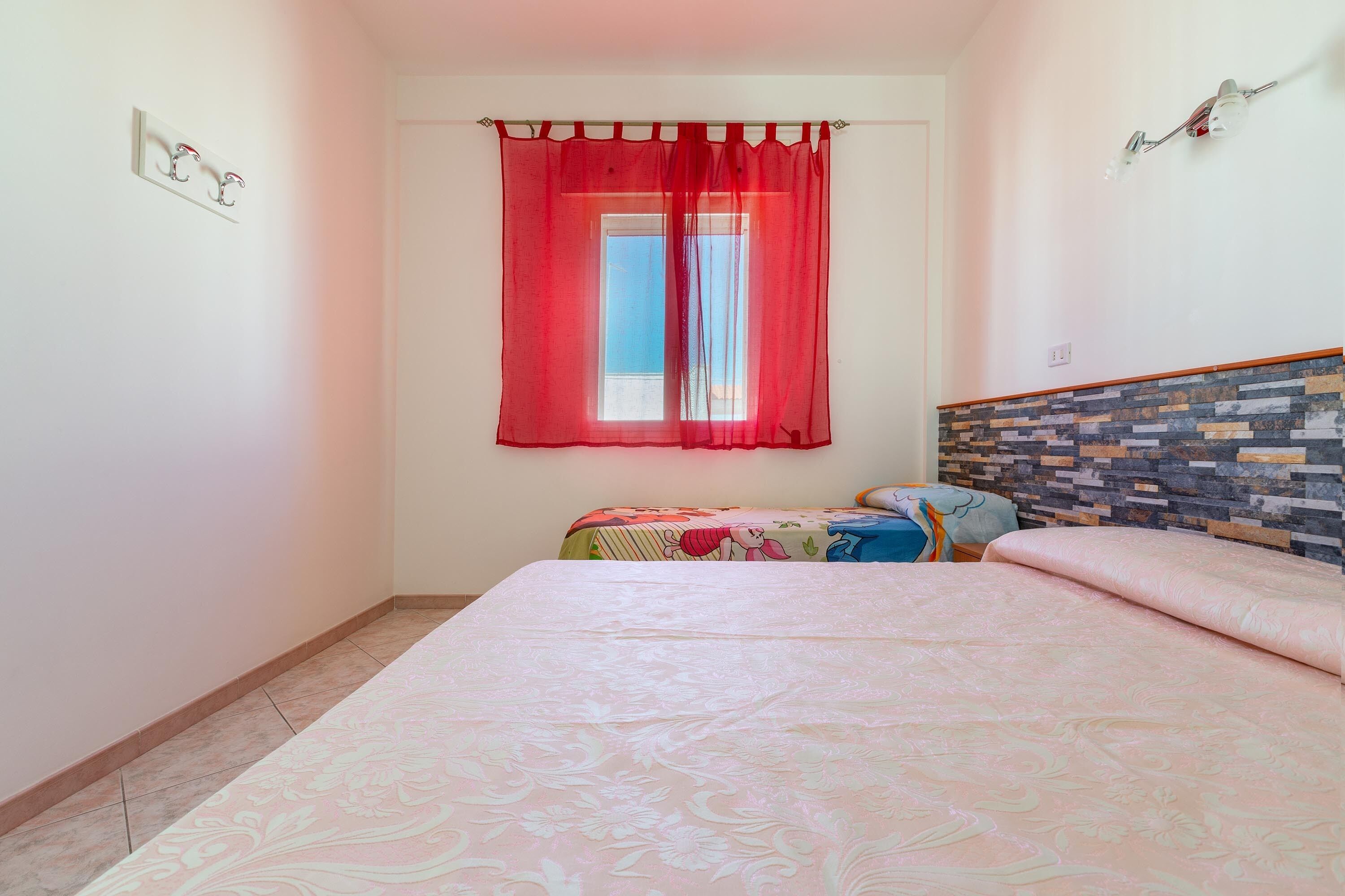 A bedroom with a bed, a window with red curtains, a wall-mounted light fixture, and a patterned headboard.