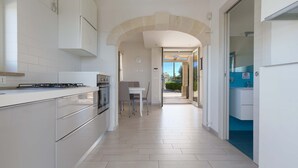 A modern kitchen with white cabinetry, a dining area with a table and chairs, and a view through an open door to a balcony. - 2097 Villa Regina Dello Ionio (Manduria)