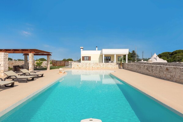 A swimming pool with lounge chairs, a pergola, and a modern house with stone walls.