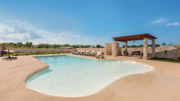 A swimming pool with a curved edge, surrounded by lounge chairs and a stone structure with a roof. - 2088 Villa Marilda (Martina Franca)
