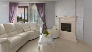 A living room with a white leather sofa, a glass coffee table with a vase of flowers, a fireplace, and a view through large windows with purple curtains. - 2088 Villa Marilda (Martina Franca)