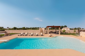 A swimming pool with clear blue water, surrounded by lounge chairs and a stone wall.