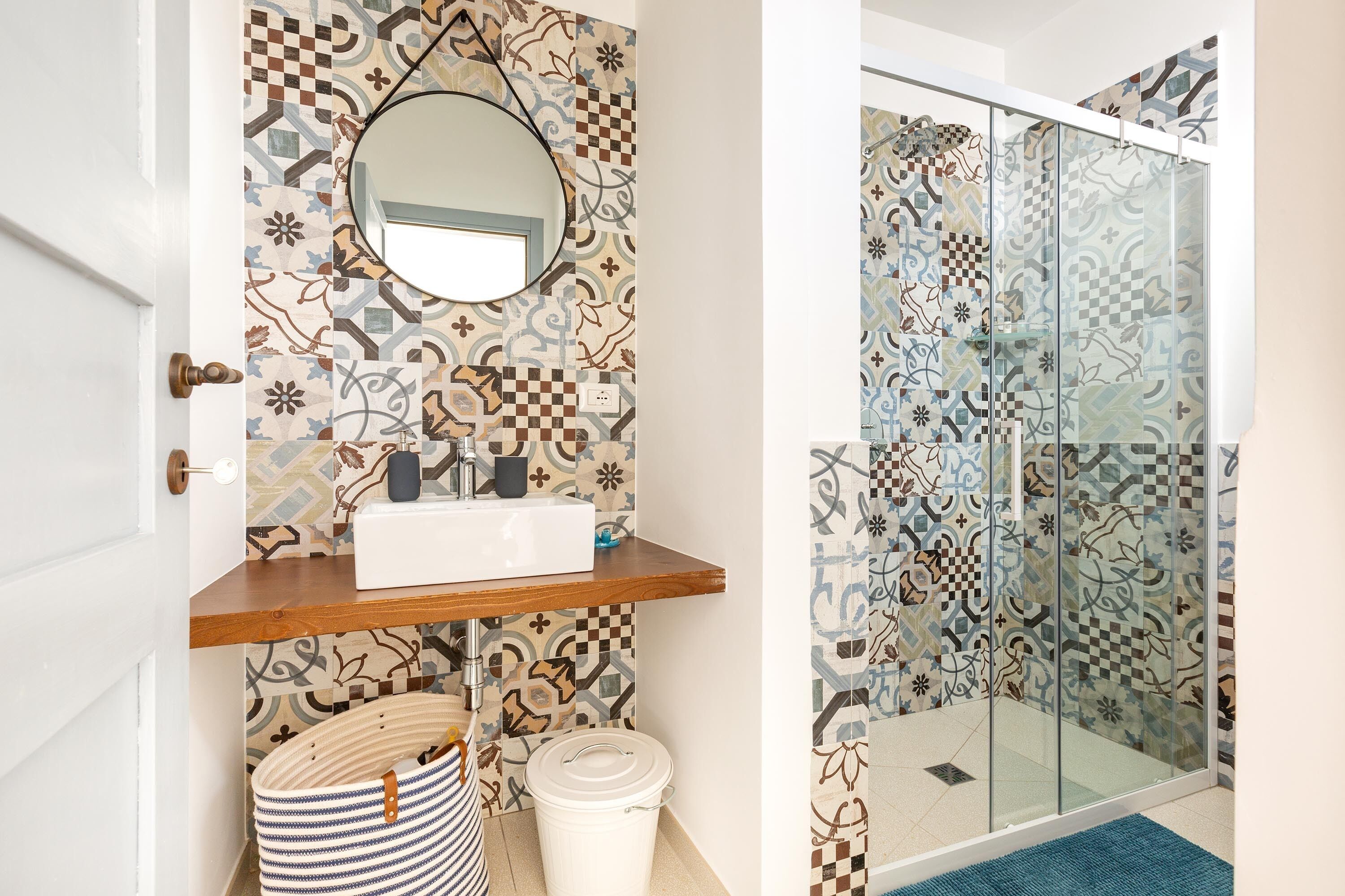 Un baño con baldosas estampadas, un estante de madera, un espejo redondo, un lavabo, un bolso a rayas y un cubo blanco.