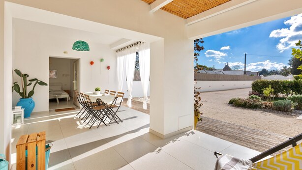 A bright, open-plan living area with a dining table and chairs, a large potted plant, and a view of a garden and building in the background. - 2124 I Colmi Trulli Suites (Martina Franca)