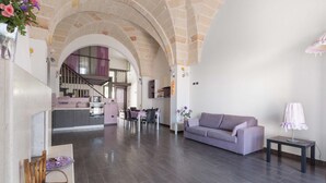 A spacious living area with a modern kitchen in the background, a purple sofa, and a small table with a lamp. - 2094 La Dolce Vita Suite (Avetrana)