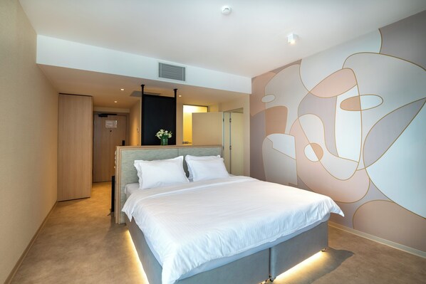 Een moderne hotelkamer met een groot bed, een nachtkastje met een bloemenvaas, een groot abstracte muurschildering en een deur naar een andere kamer.