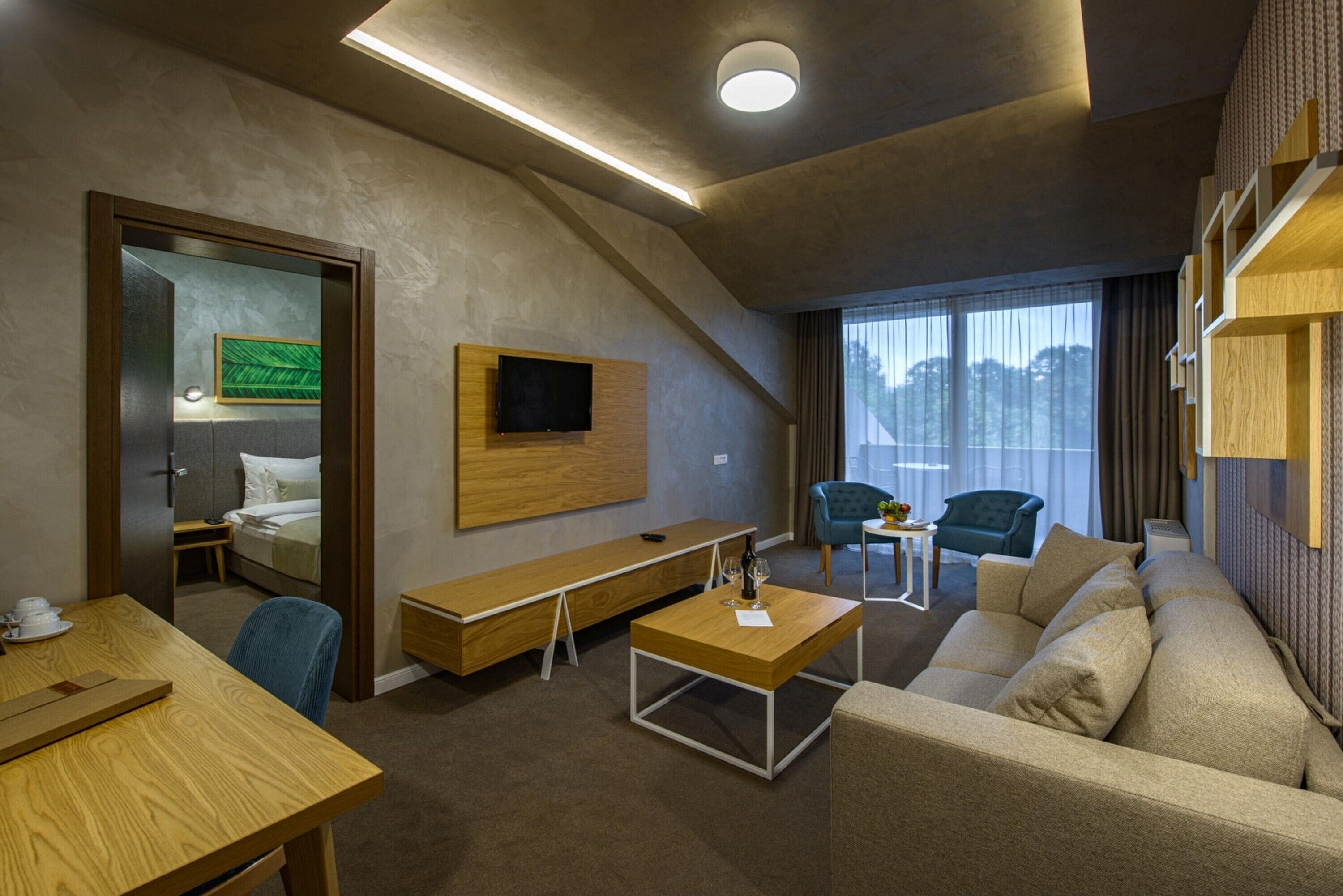A modern hotel room with a bed, a wooden desk, a sofa, a TV mounted on the wall, and a large window with a view.