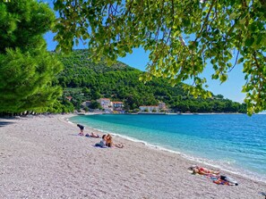 Une plage de galets où des gens se prélassent au soleil, une mer d’un bleu limpide et, au loin, des bâtiments sur une colline.