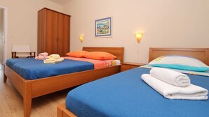 Une chambre d’hôtel avec deux lits en bois, chacun agrémenté de draps et d’oreillers bleus. On y trouve des serviettes sur les lits, une armoire en bois, un tableau au mur et deux lampes fixées au mur.