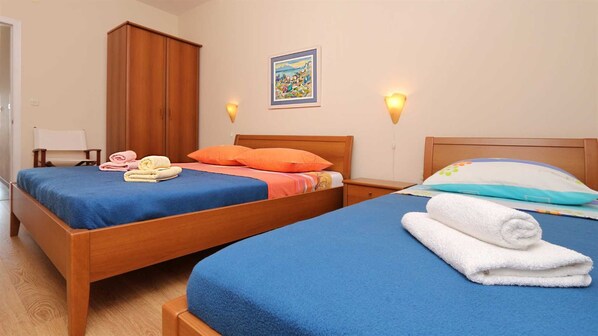 A hotel room with two wooden beds, each with blue bedding and pillows. There are towels on the beds, a wooden wardrobe, a painting on the wall, and two wall-mounted lights. - Sea View Apartment 23 (Drvenik)