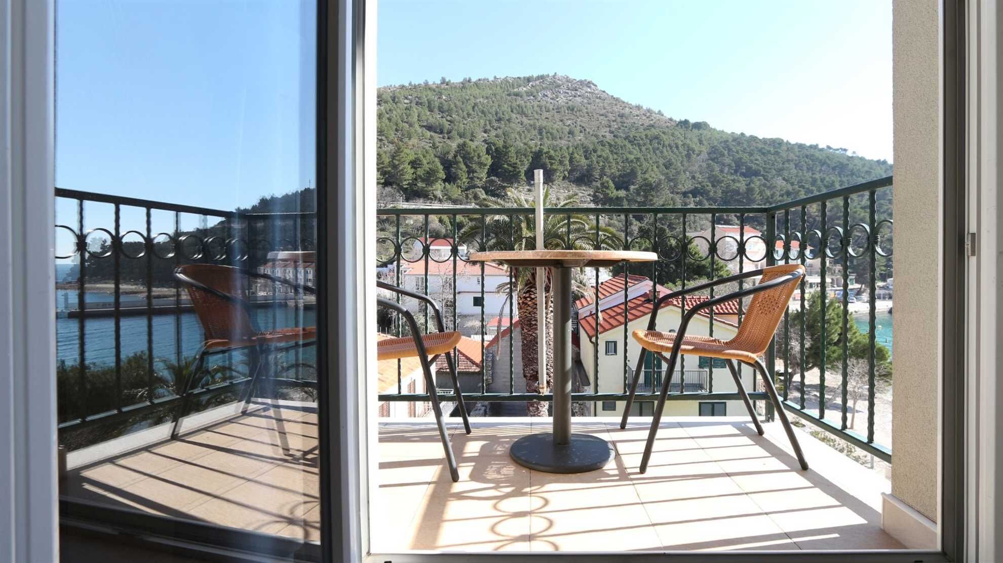 A balcony with a table and two chairs, overlooking a coastal area with a hill in the background.