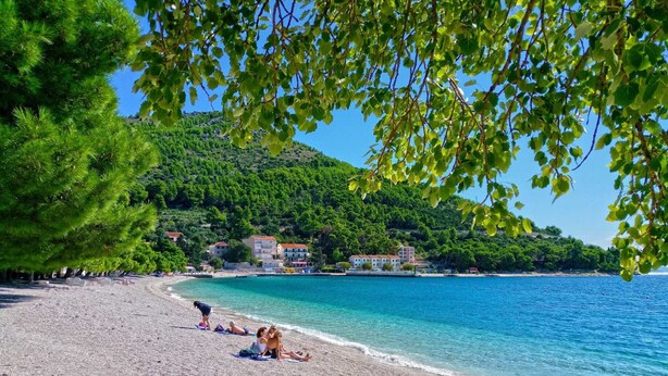 A pebbled beach with people sunbathing, a clear blue sea, and buildings on a hillside in the background. - Sea View Apartment 33 (Drvenik)