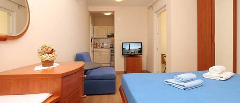 A hotel room with a bed, a desk, a chair, a TV, and a kitchenette in the background.