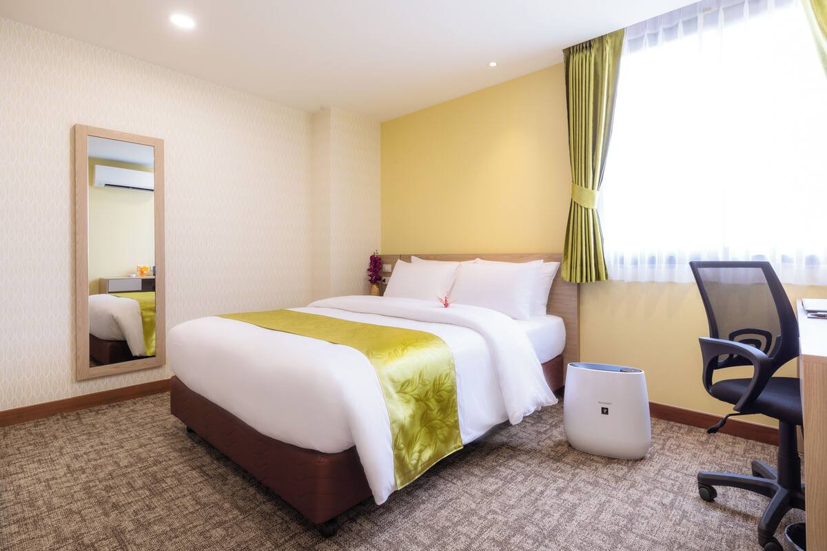 Une chambre d’hôtel avec un lit, un bureau, une chaise, un miroir et une fenêtre avec des rideaux.
