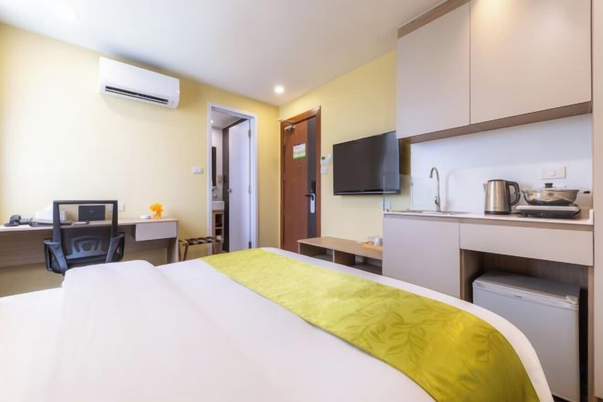 A hotel room with a bed, desk, chair, kitchenette, and a TV.
