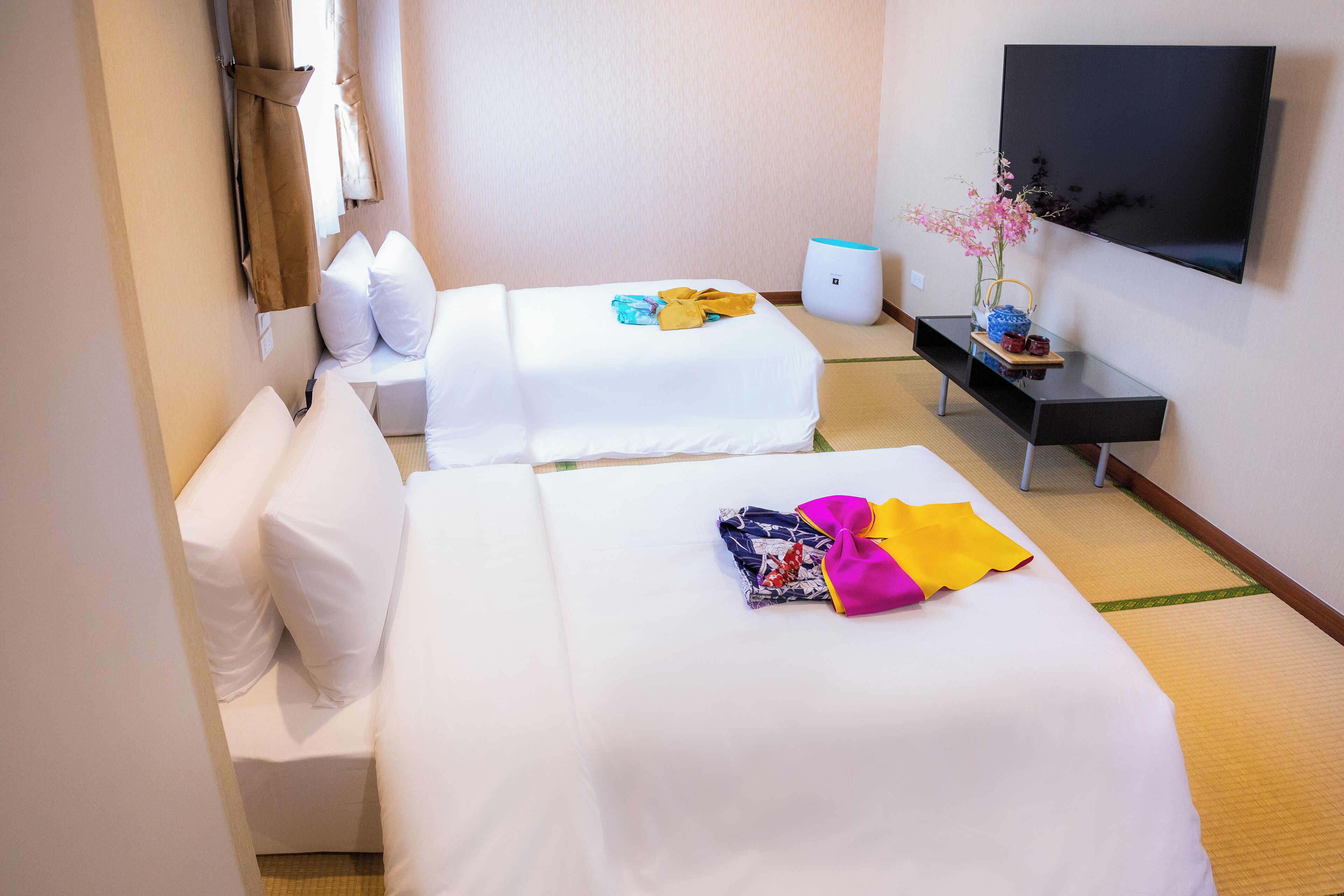 A hotel room with two beds, a TV, a small table with a vase of flowers, and a wall-mounted TV.