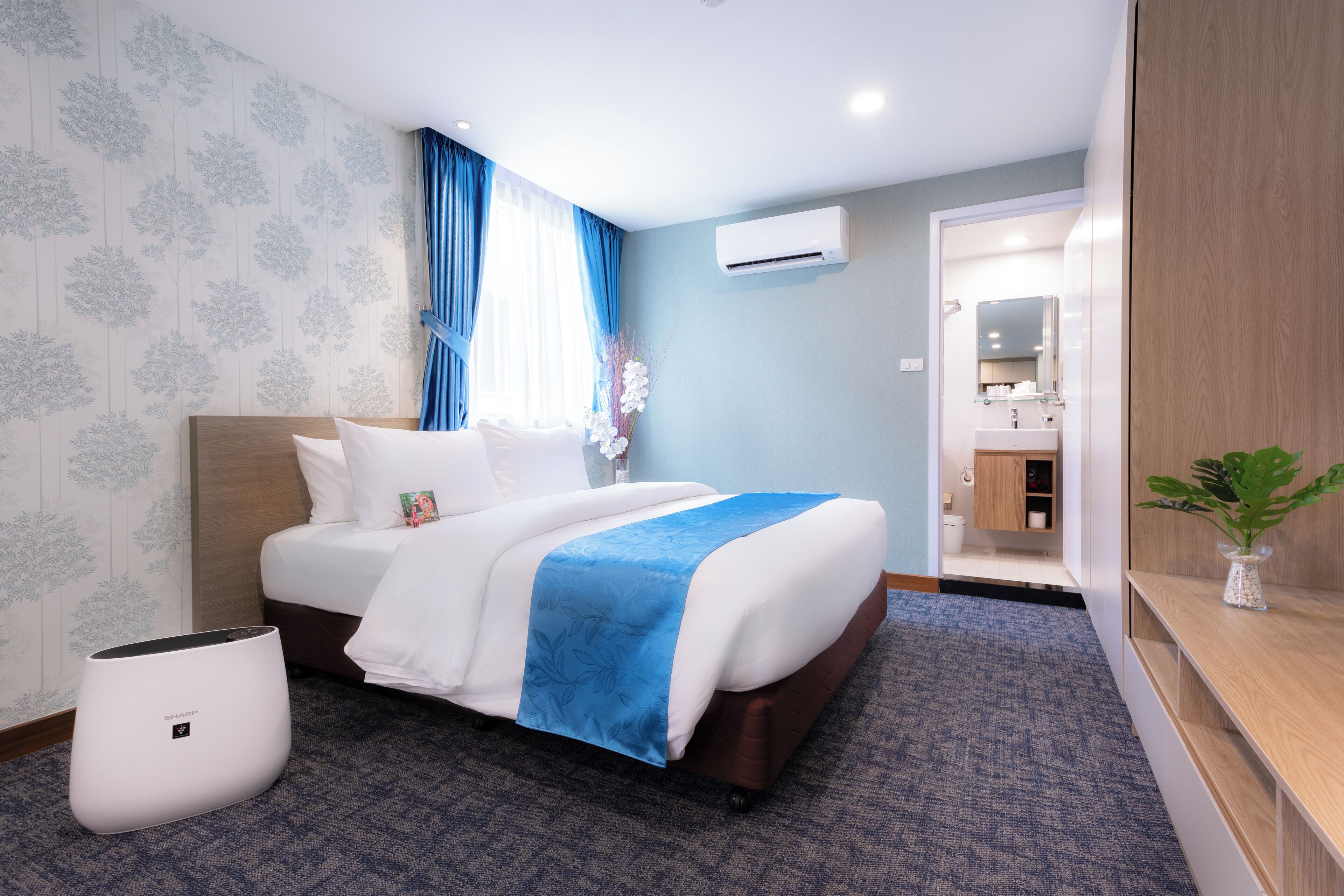 Een hotelkamer met een bed, een blauw-witte bedspreider, een witte airconditioner, een blauw tapijt en een houten nachtkastje met een bloevas.