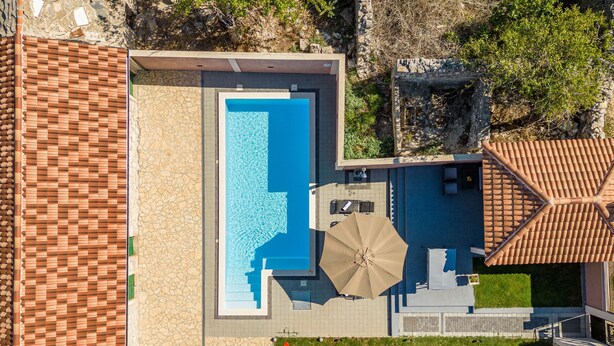 Luchtbeeld van een modern huis met een rechthoekig zwembad, een terras met tafel en parasol, en een stenen muur.