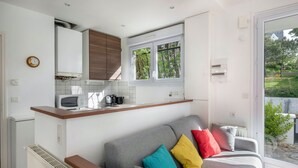 A compact living space with a kitchenette, a grey sofa with colorful cushions, and a view of greenery outside. - 200m from the beach, cosy house for 4 (La Baule-Escoublac)