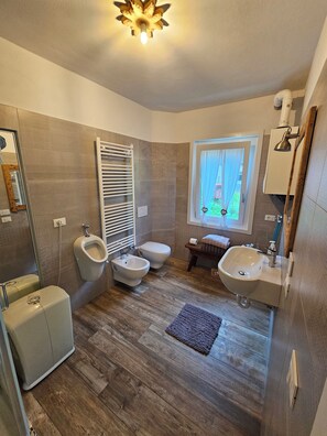 A modern bathroom with a bidet, toilet, and sink, wooden flooring, and a window with curtains.