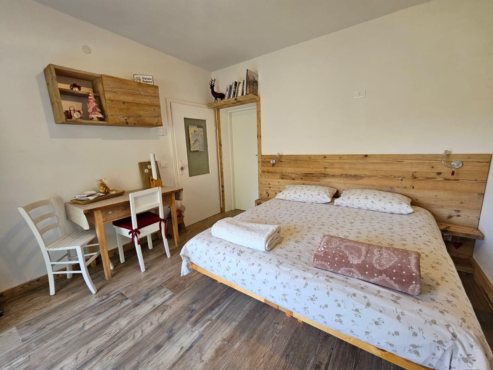 A bedroom with a wooden bed, a desk with a chair, and a small shelf with decorative items.