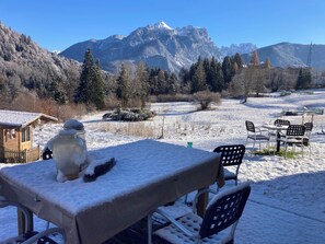 A snow-covered outdoor dining area with a table and chairs, a wooden cabin, and a mountainous backdrop.