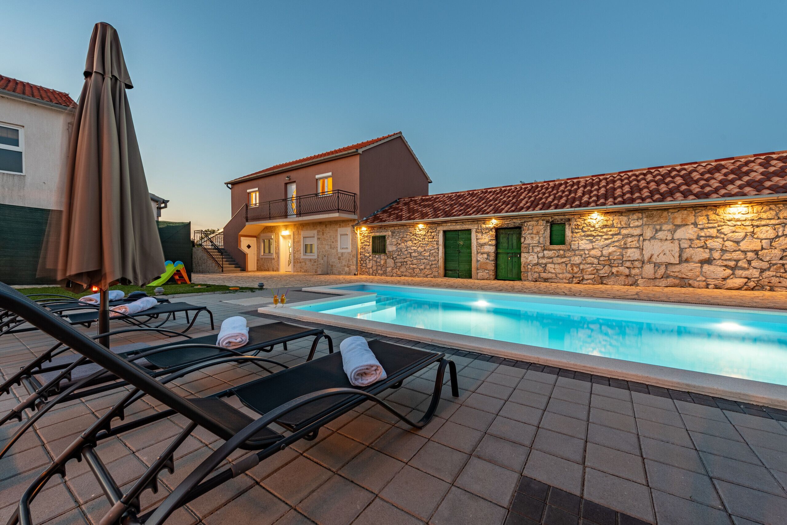 A swimming pool area with lounge chairs and a stone building with a tiled roof.