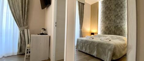 A hotel room with a bed, a desk, a chair, a flat-screen TV mounted on the wall, and a window with curtains.