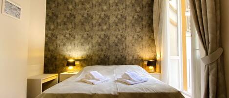 Une chambre d’hôtel avec un grand lit, deux tables de chevet avec des lampes, un mur décoré d’un motif et une fenêtre avec des rideaux.