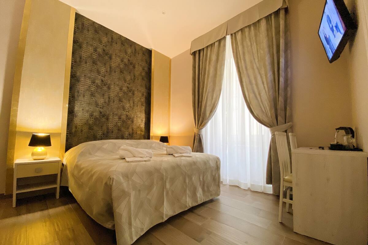 A hotel room with a large bed, two bedside tables with lamps, a desk with a chair, a television mounted on the wall, and a window with curtains.