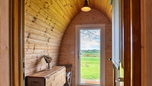 L’intérieur d’un chalet accueillant, avec des murs en bois, un banc, une petite table ornée d’un vase et une porte donnant sur un espace extérieur verdoyant.
