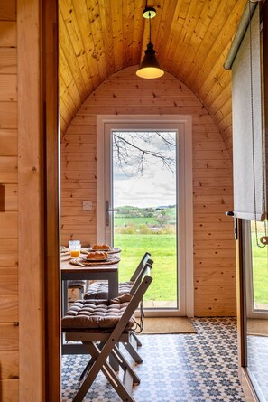 A cozy wooden cabin with a curved ceiling, a dining table with pastries and a glass of juice, and a view of a green landscape through a large door. - The Pods (Camerton)