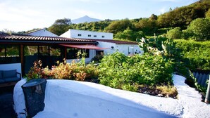 A white building with a red roof, surrounded by greenery and a mountain in the background.