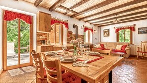 A dining area with a wooden table, chairs, and a sofa. There is a kitchen area with a stove and cabinets. The room has a wooden ceiling and large windows. - House By The Stream in Soca Valley (Kanal)