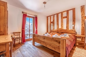 A wooden bed with a headboard, a wooden desk, and a chair in a room with a view of a snowy landscape through a window.