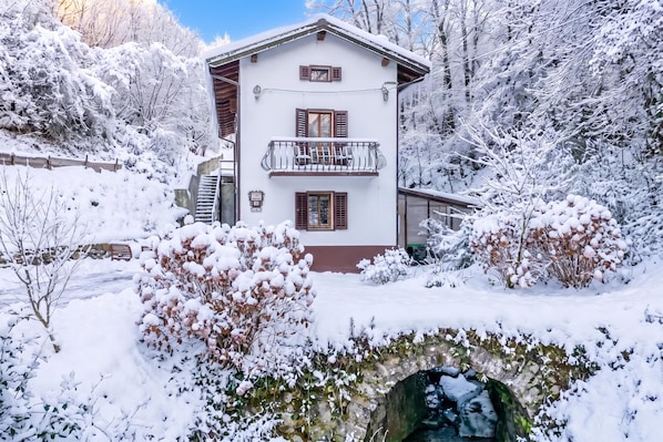 A two-story chalet with a balcony, surrounded by snow-covered trees and a stone bridge.