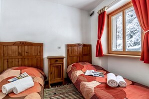 A bedroom with two wooden beds, a small wooden nightstand, a window with red curtains, and a view of snow-covered trees.