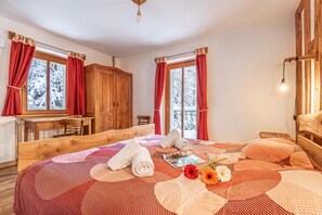A cozy bedroom with a wooden bed, a desk, and a view of snowy trees through the windows. - House By The Stream in Soca Valley (Kanal)