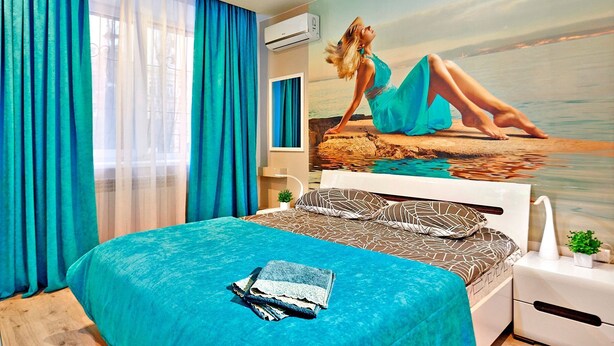 Une chambre à coucher avec un lit, une fresque représentant une femme au bord de la mer et une fenêtre avec des rideaux bleus.