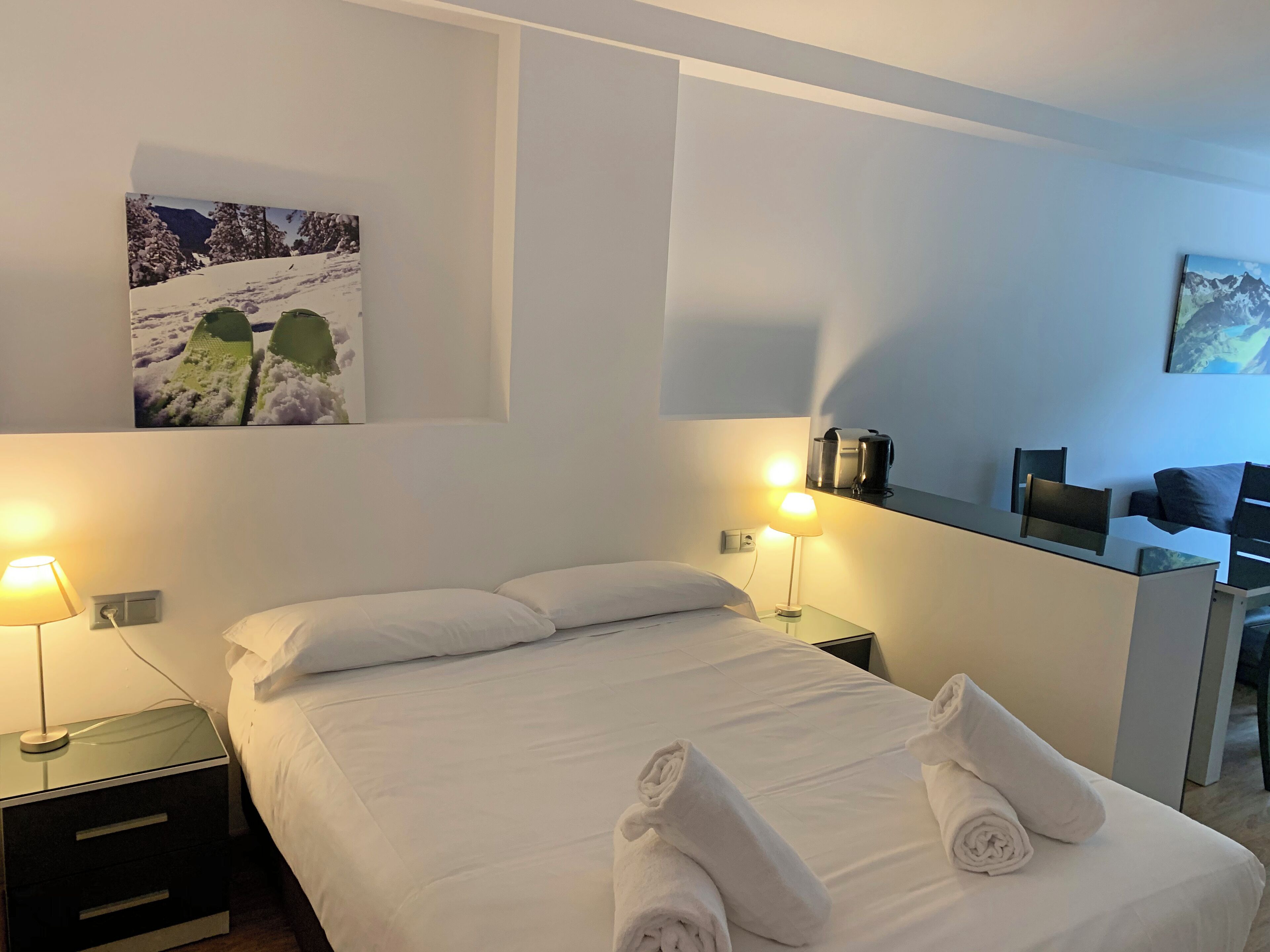 A hotel room with a bed, two bedside lamps, a nightstand, a desk with a chair, and a coffee machine.