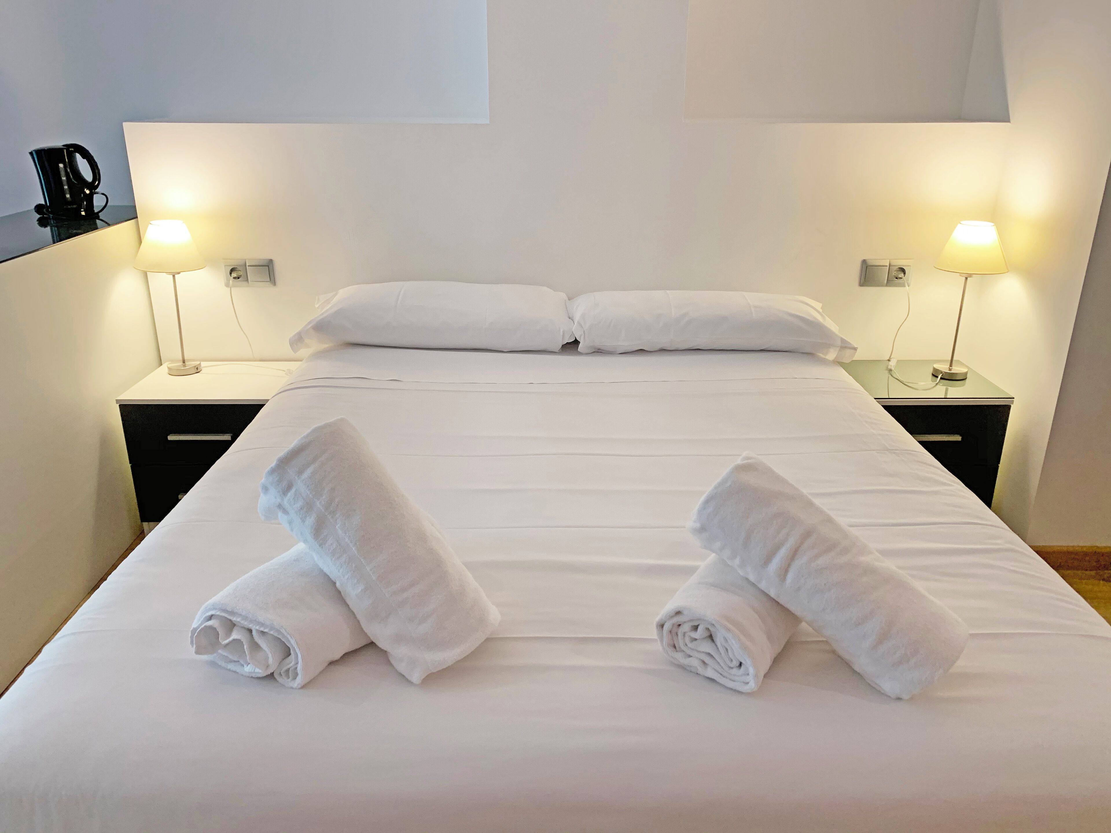 Een net opgemaakt bed met twee opgerolde handdoeken ervoor, twee bedlampjes en een zwarte koffiezetapparaat op het nachtkastje.
