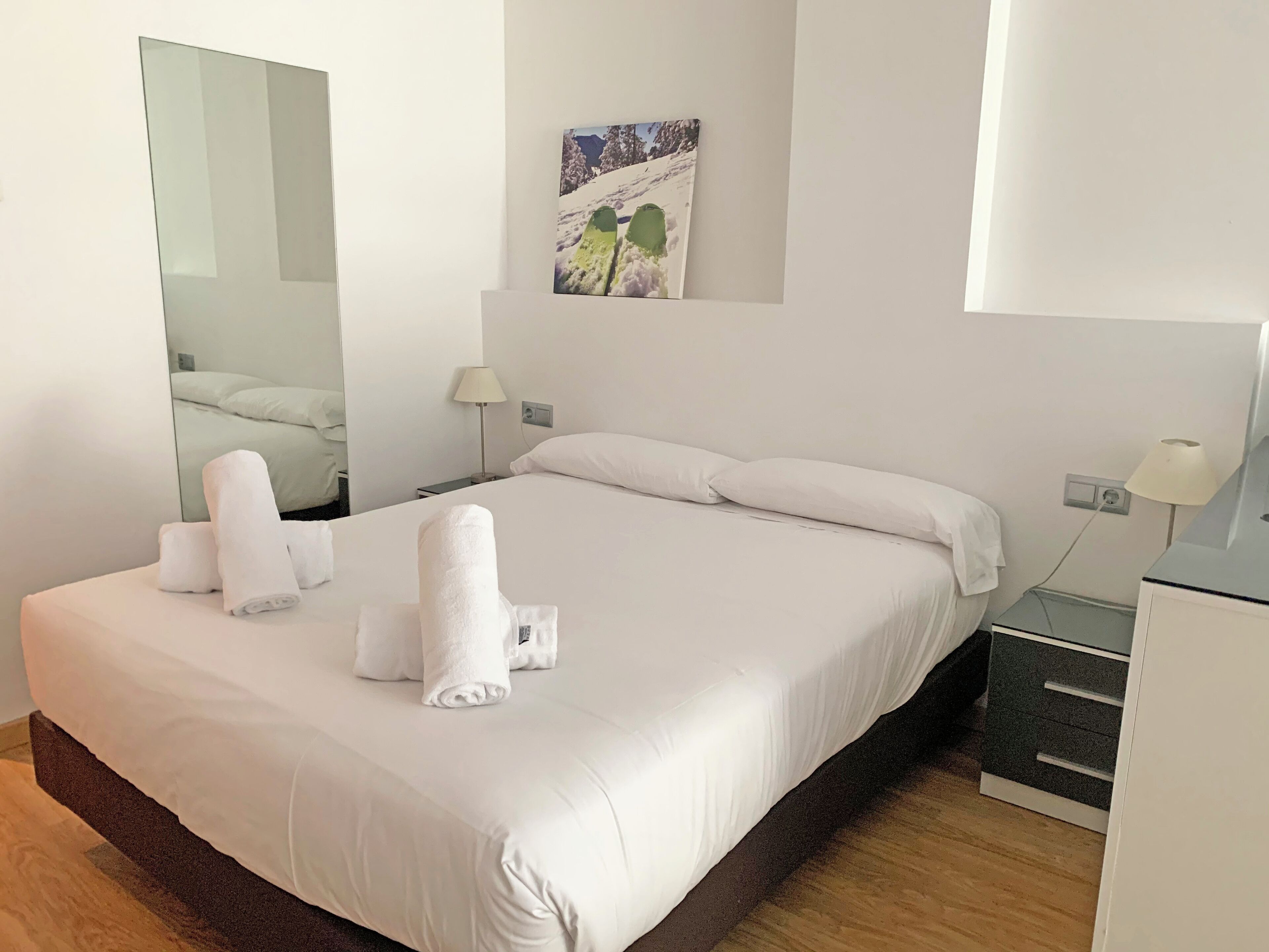 A hotel room with a large bed, white linens, a mirror, a painting, and a desk with a lamp.