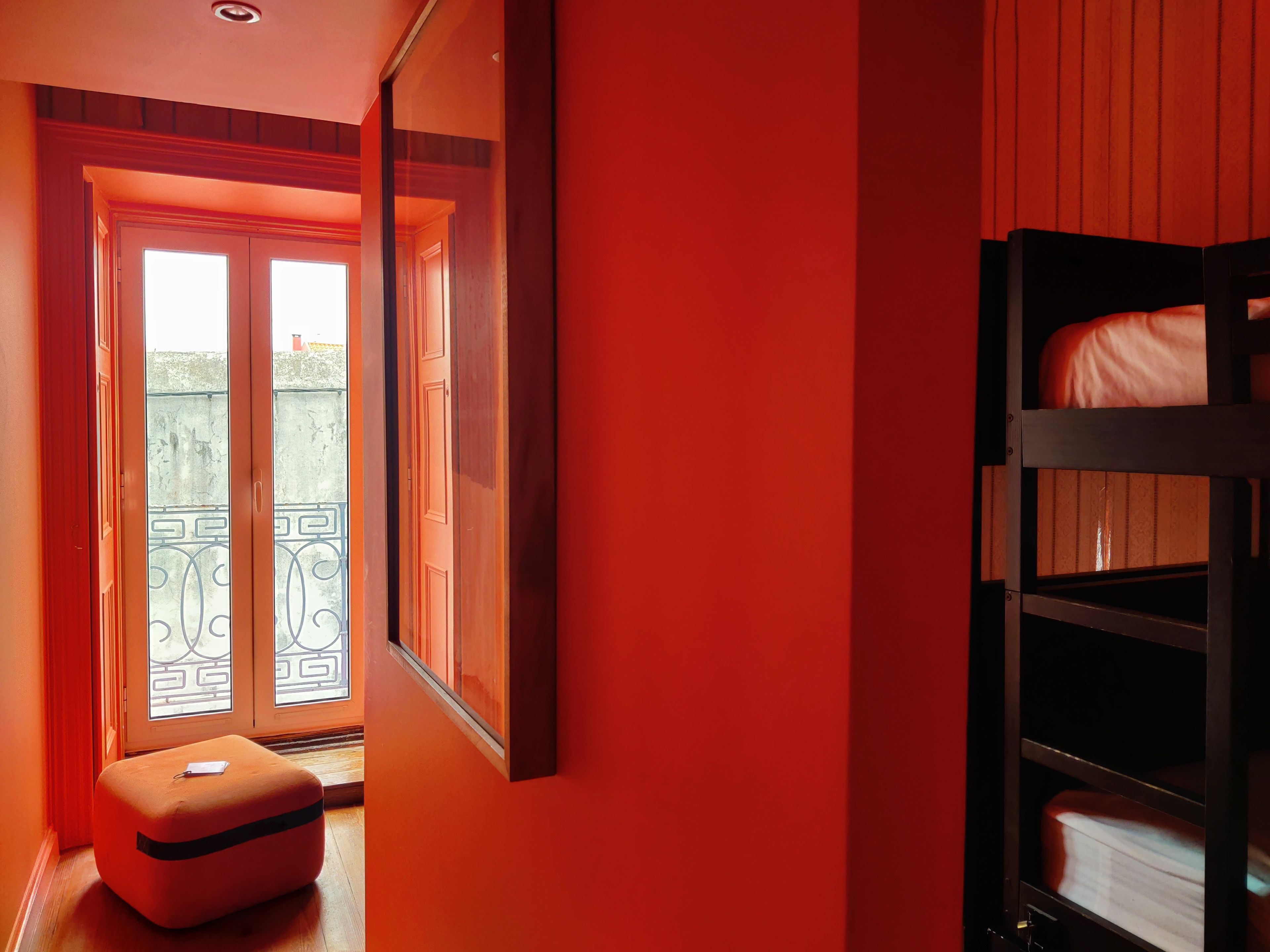 Une pièce avec un mur rouge, un banc en bois, un lit superposé et une porte donnant sur un balcon agrémenté d’une rambarde en métal.