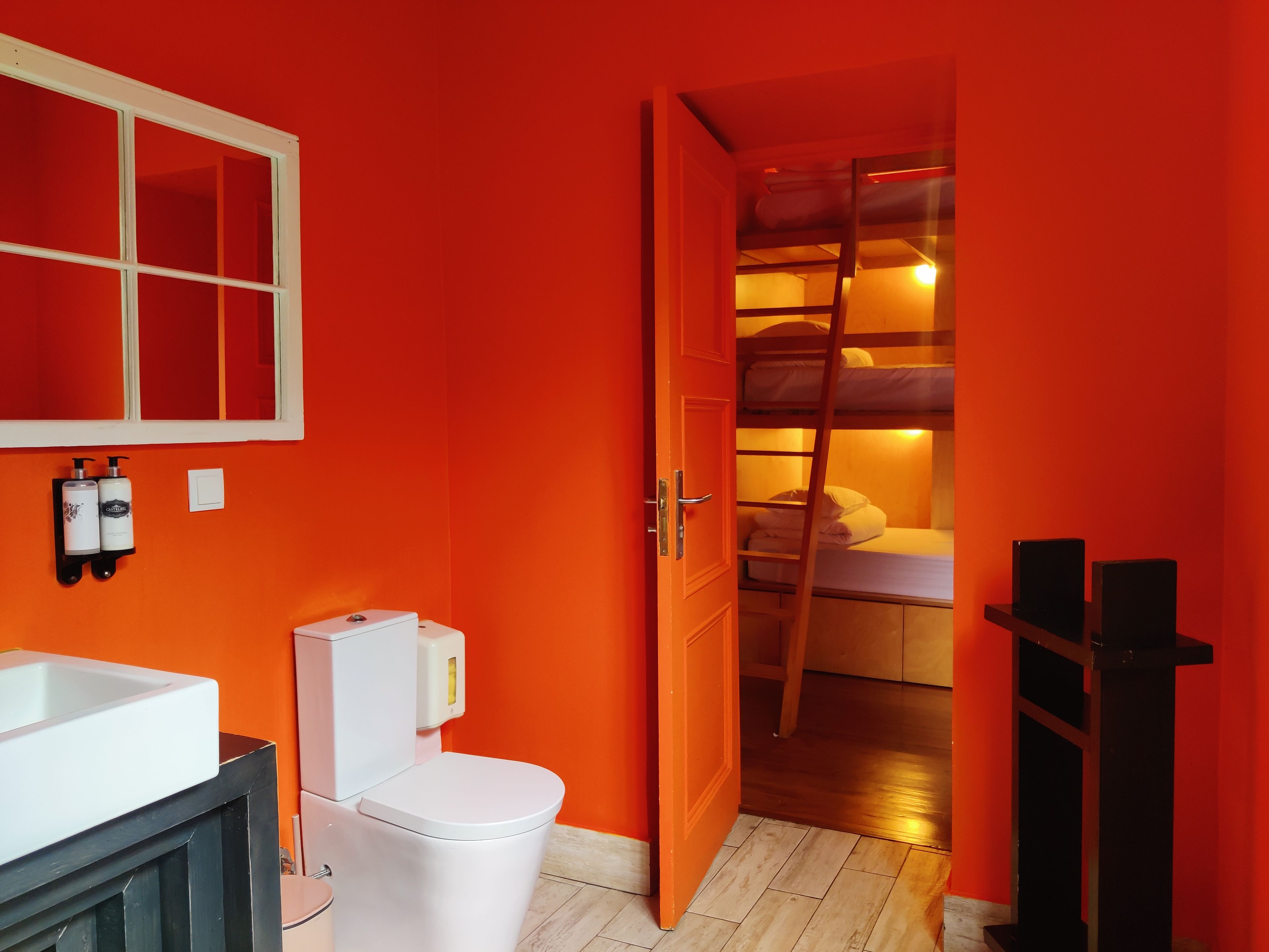 Un baño con un lavabo blanco, inodoro y espejo. La pared es roja y el suelo de madera.