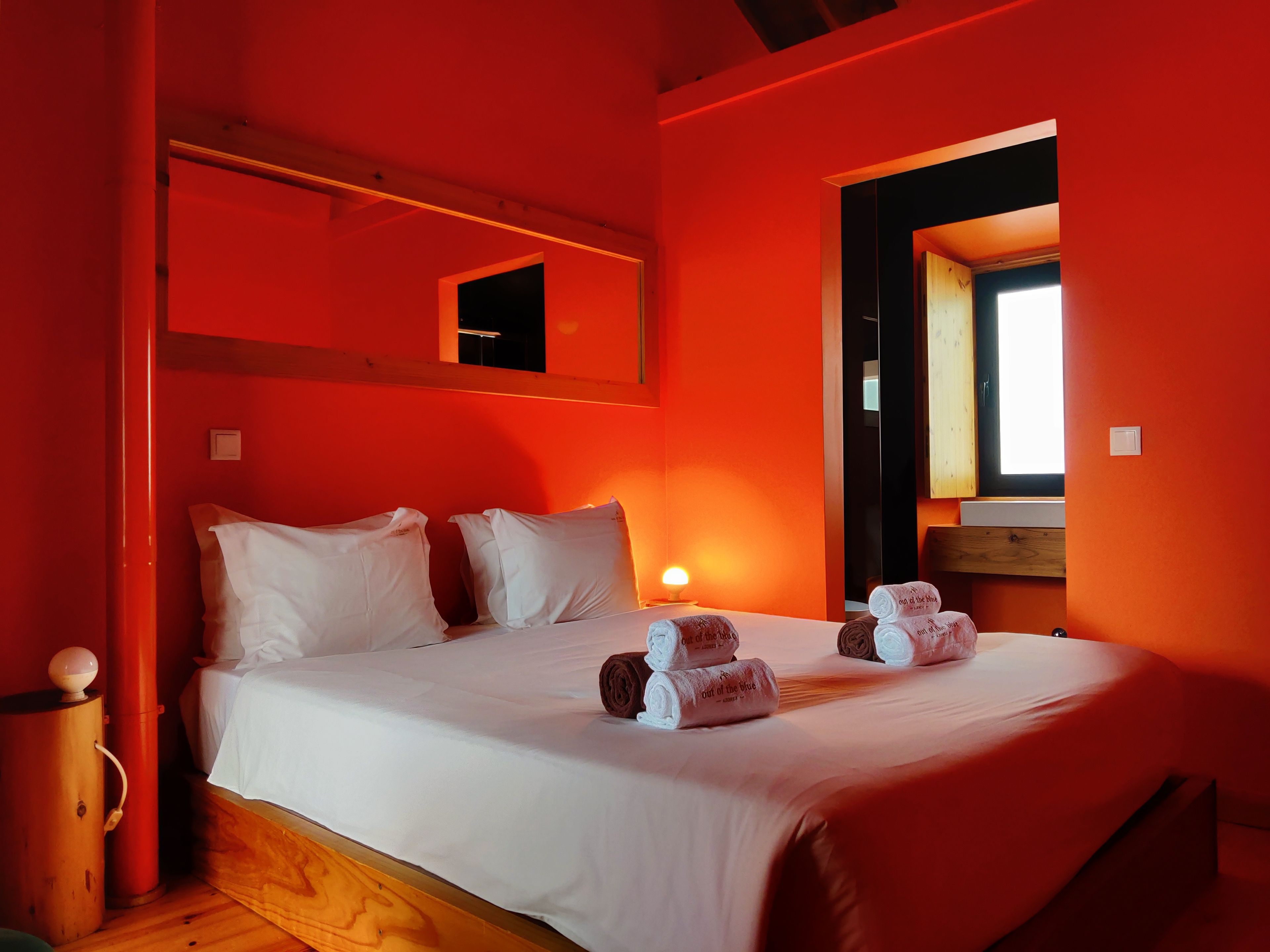 A room with a bed, pillows, a bedside lamp, a wooden floor, a red wall, and a window.