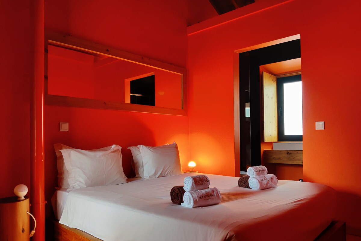 A room with a bed, pillows, a bedside lamp, a wooden floor, a red wall, and a window.