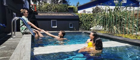 Una piscina con cubierta de madera y barandillas de metal, rodeada de vegetación y edificios.